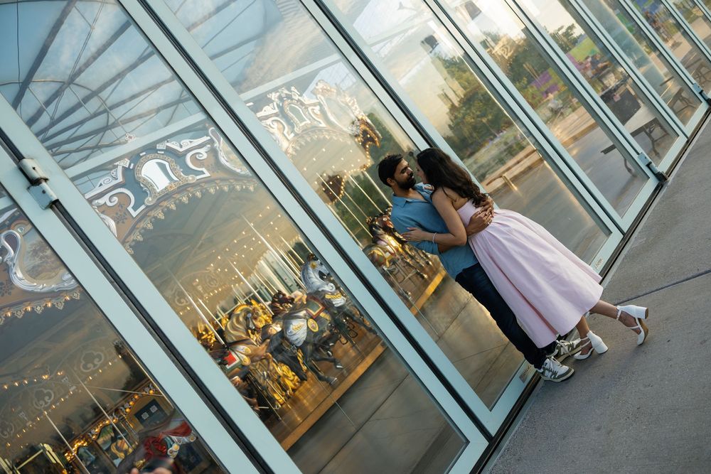 Couple sharing a romantic kiss outside a carousel, woman in pink skirt and man in blue shirt, reflecting in glass windows.
