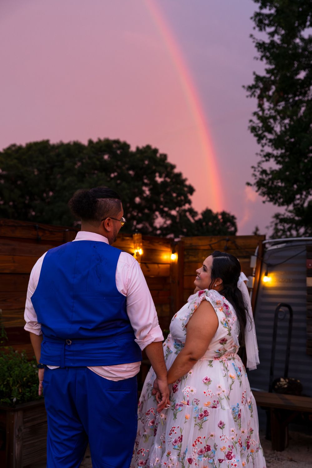 A couple in formal attire views a vibrant rainbow at sunset while standing in a garden setting.