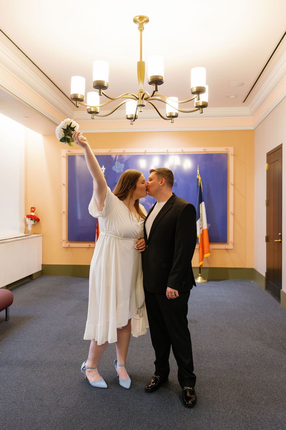 Couple sharing a kiss at city hall wedding ceremony, bride in white dress holding bouquet aloft, groom in black suit.
