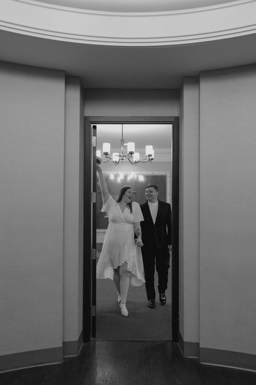 Black and white photo of a joyful couple walking through an elegant doorway, woman in white dress raising her arm in celebration.