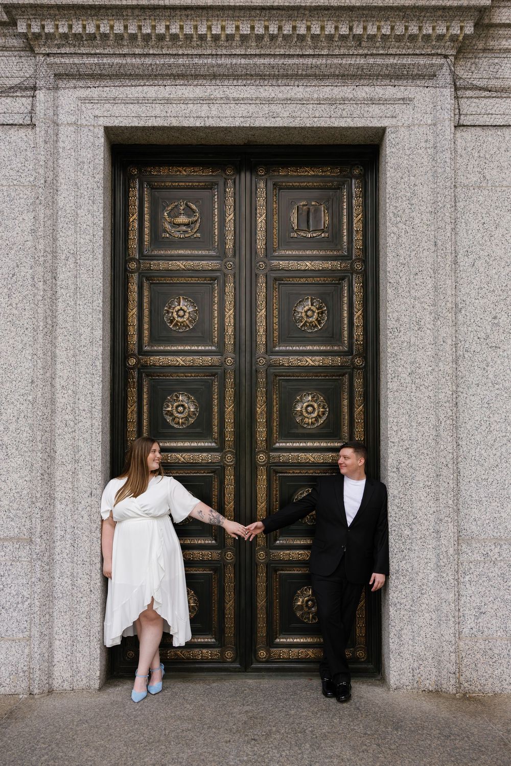 Couple holding hands in front of ornate bronze doors, woman in white dress and blue heels, man in black suit.