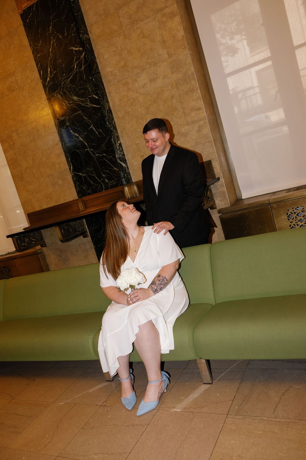 Bride in white dress and blue heels sits on green sofa laughing with groom in black suit at wedding venue.