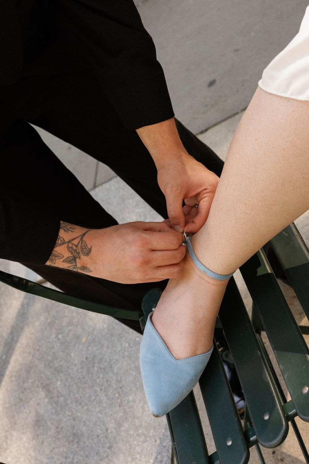 Tattooed hands fastening a blue ankle strap on a light blue pointed-toe flat shoe while seated on a bench.