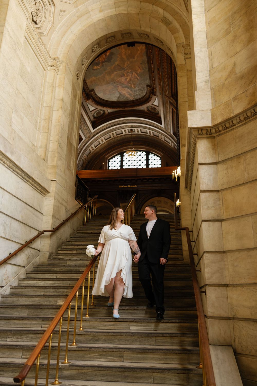 Bride and groom walk down grand stone staircase in ornate historic building, bride holding white bouquet.