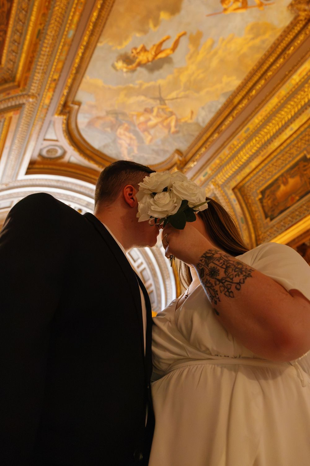 Couple sharing a kiss at their wedding under an ornate gold ceiling with classical fresco paintings.