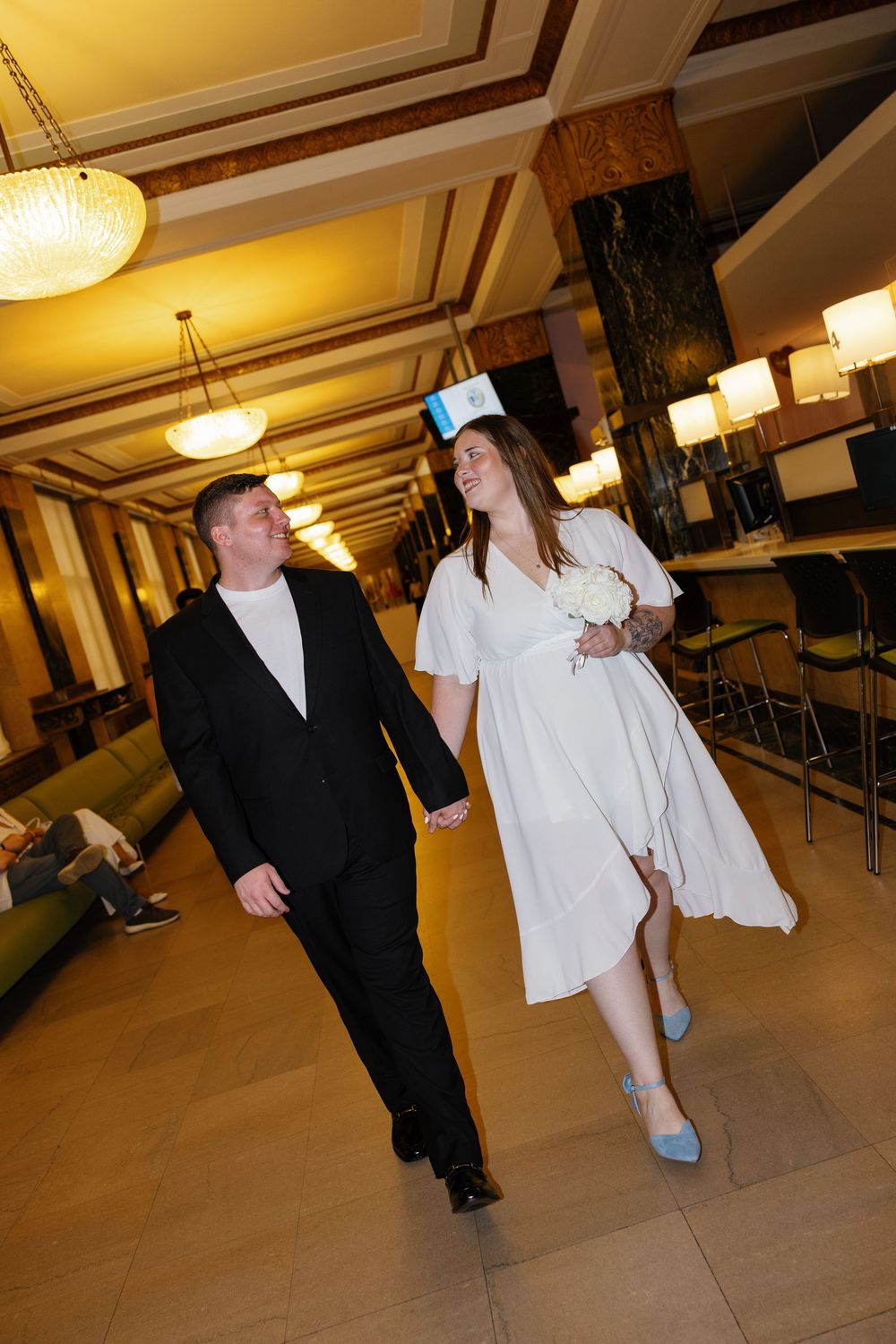 Bride in white dress and blue heels holds bouquet while walking hand-in-hand with groom in black suit through elegant hotel lobby.