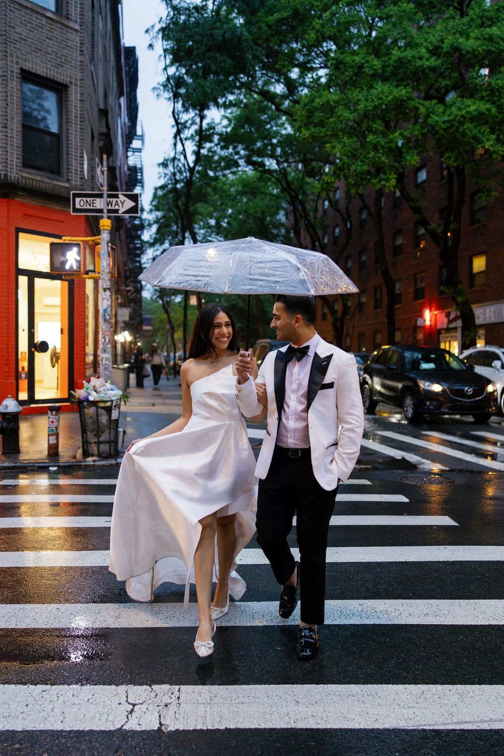 Bride and groom cross a rainy NYC street under a clear umbrella, dressed in elegant wedding attire.