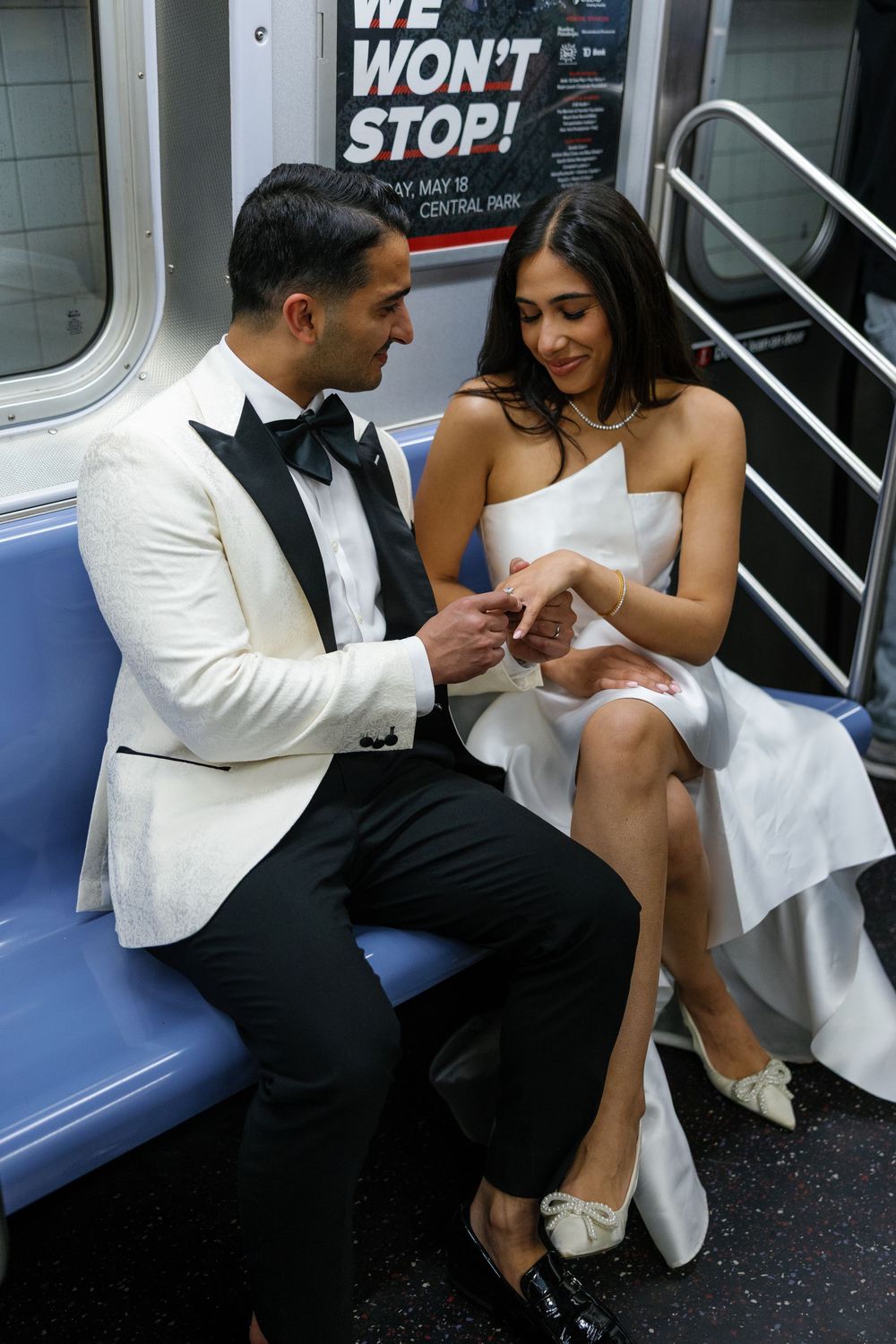A couple in formal attire sits together on a subway train, with the person in white examining something in their hands.