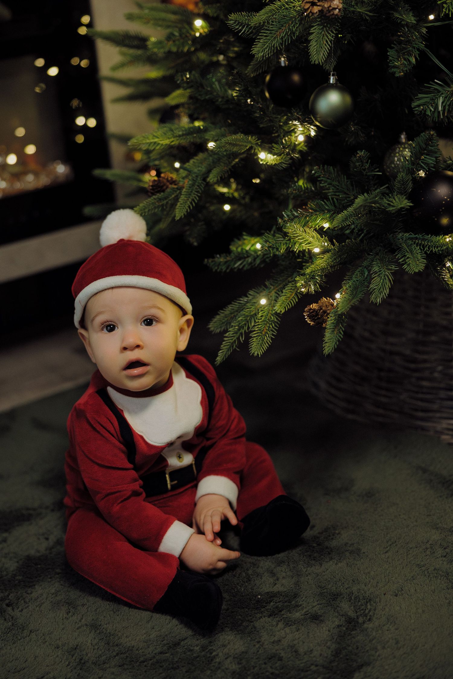 Small child in red holiday outfit crawls on carpet beneath decorated Christmas tree with twinkling lights.