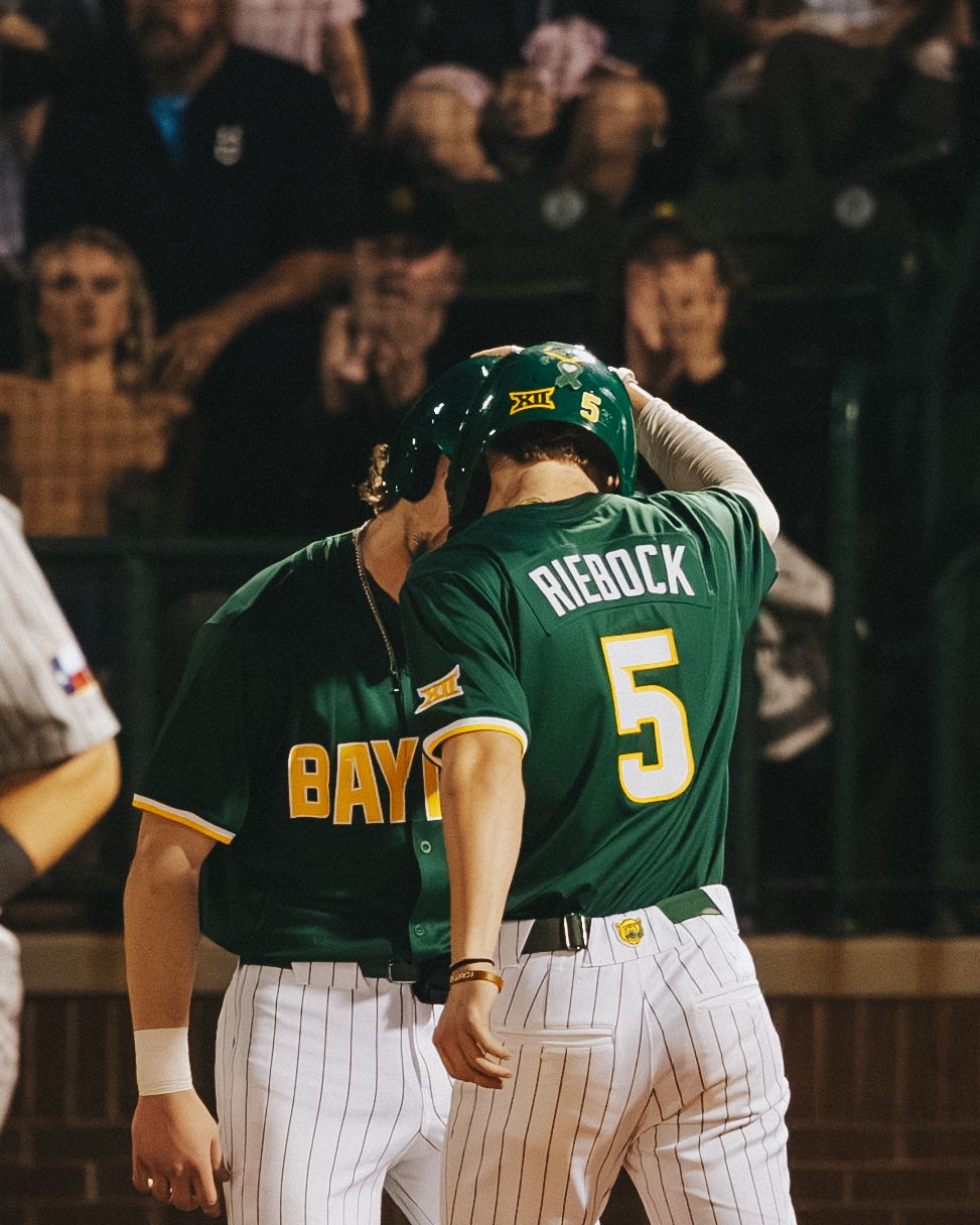 Two Baylor Bears baseball players in green uniforms celebrate together on the field, with player Rienock wearing number 5.