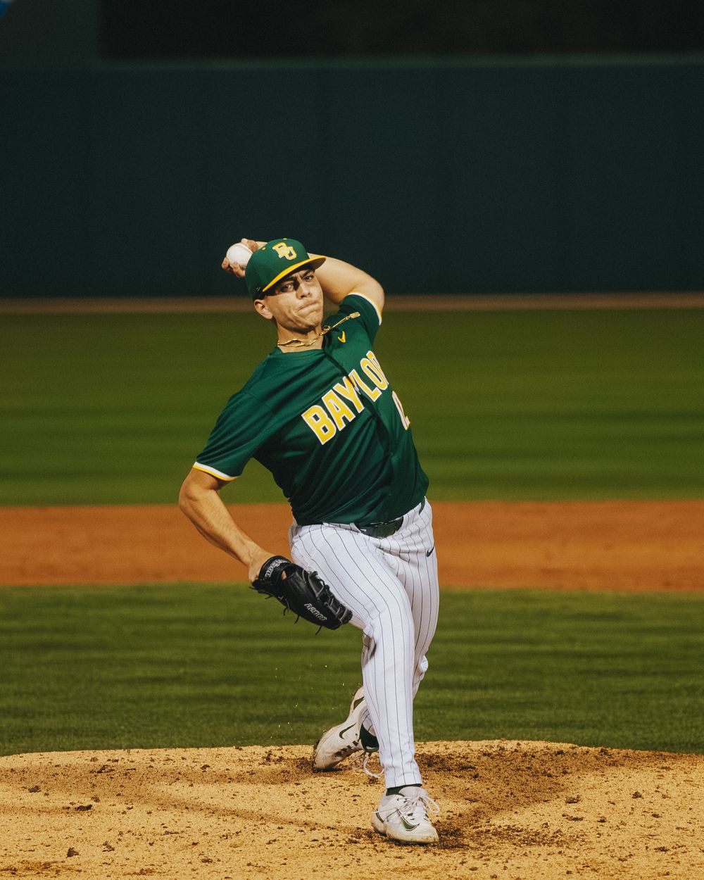 Baylor Bears pitcher winds up to deliver a pitch during a night game wearing green and white pinstripe uniform.