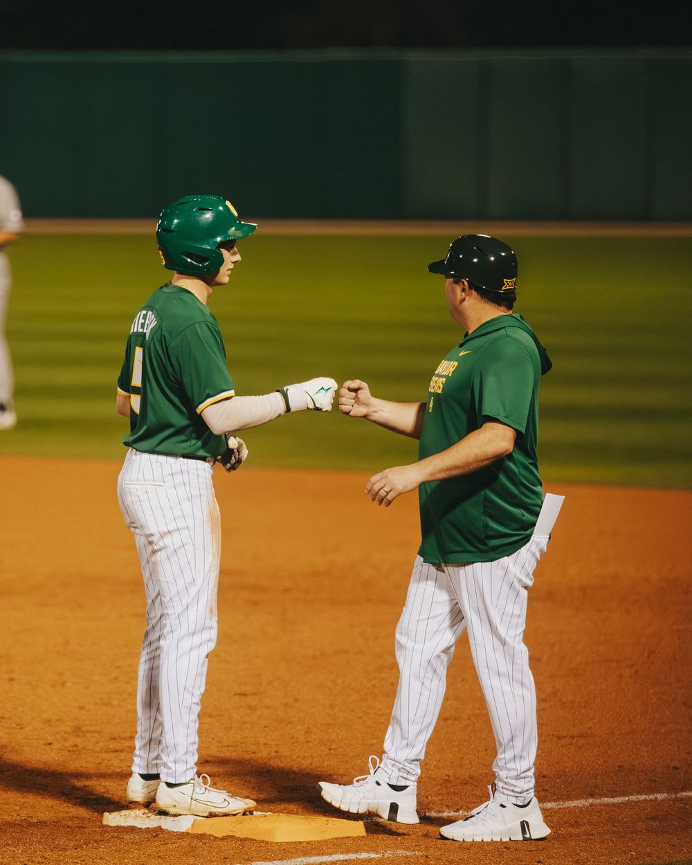 Two Baylor Bears baseball players share a fist bump at first base during a college baseball game.
