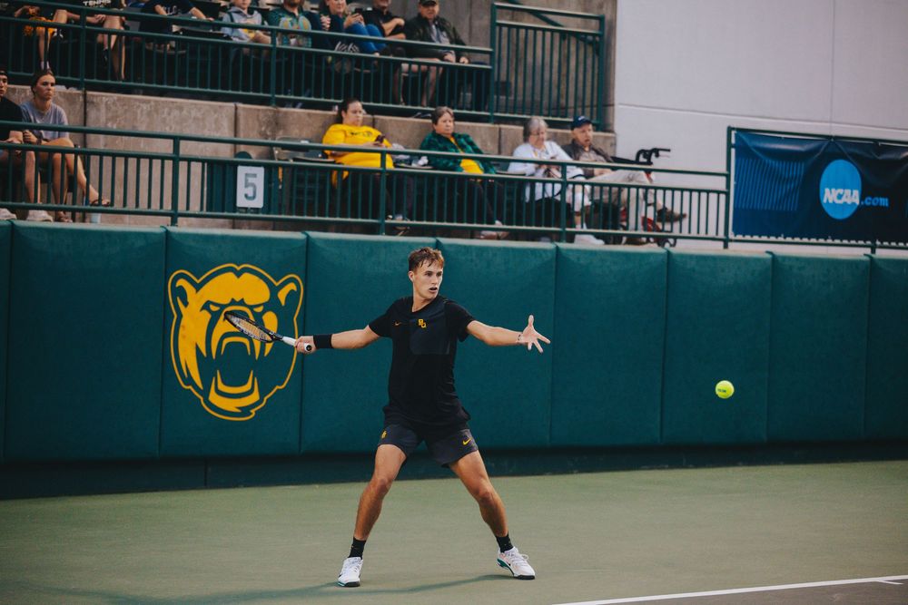 A Baylor University Men's Tennis player prepares to return a shot on an indoor NCAA court.
