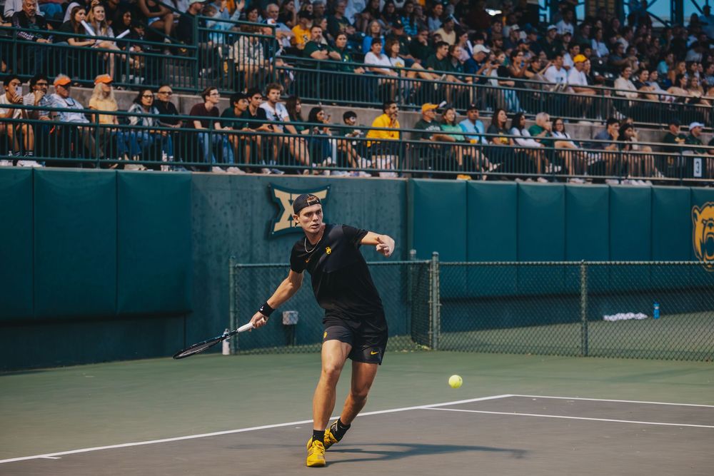 A Baylor University Men's Tennis player lunges to hit a forehand shot before a packed crowd.