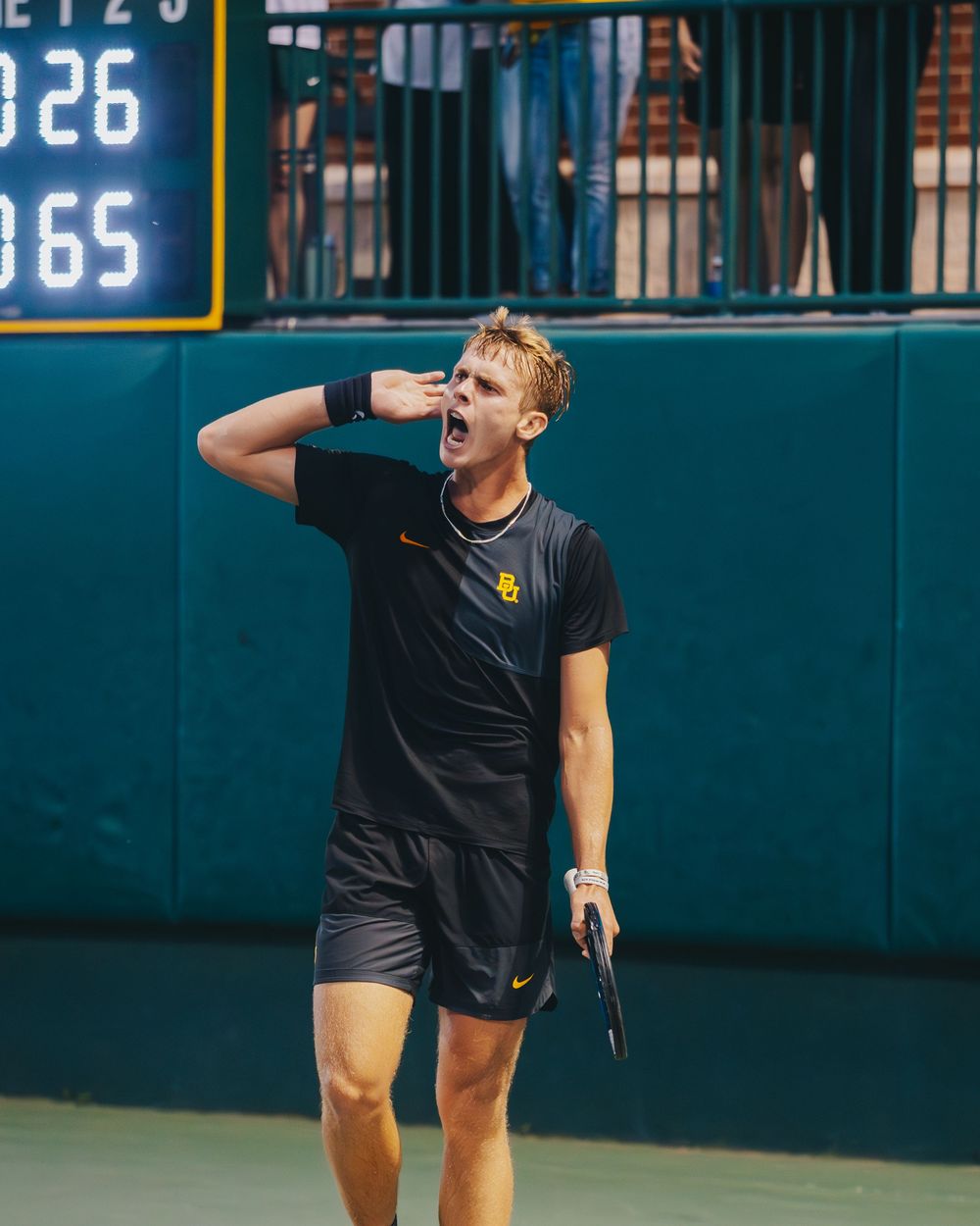 An emotional Baylor tennis player reacts passionately during a match near a scoreboard.