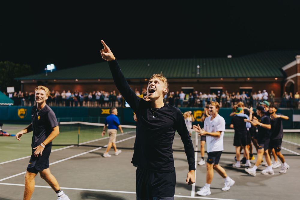 A Baylor University Men's Tennis player celebrates triumphantly on court while teammates cheer in the background.