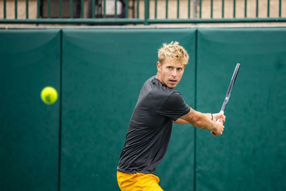 Blonde tennis player in gray shirt hits backhand shot on outdoor green tennis court.