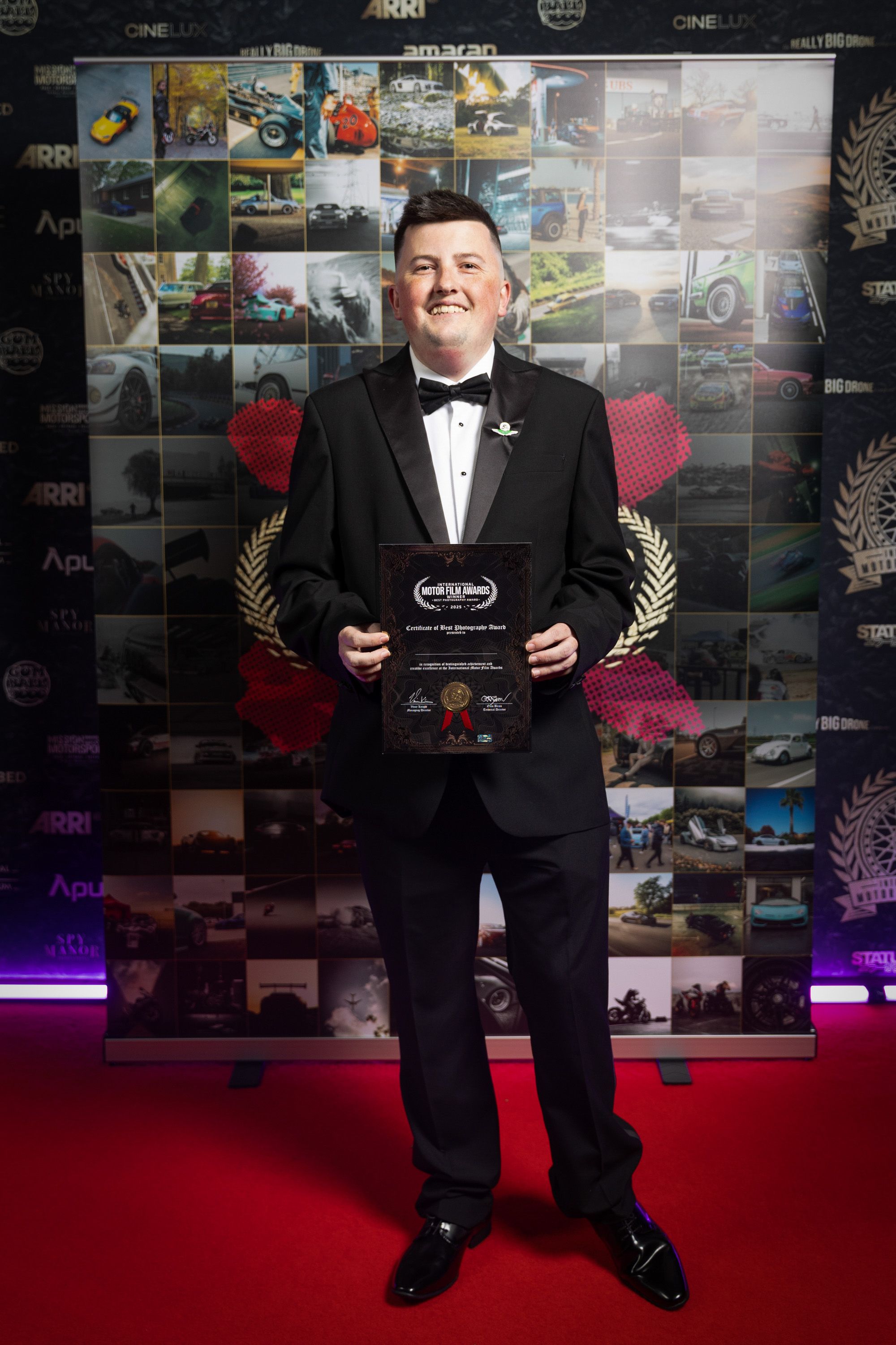 Award winner standing against photo collage wall on red carpet.