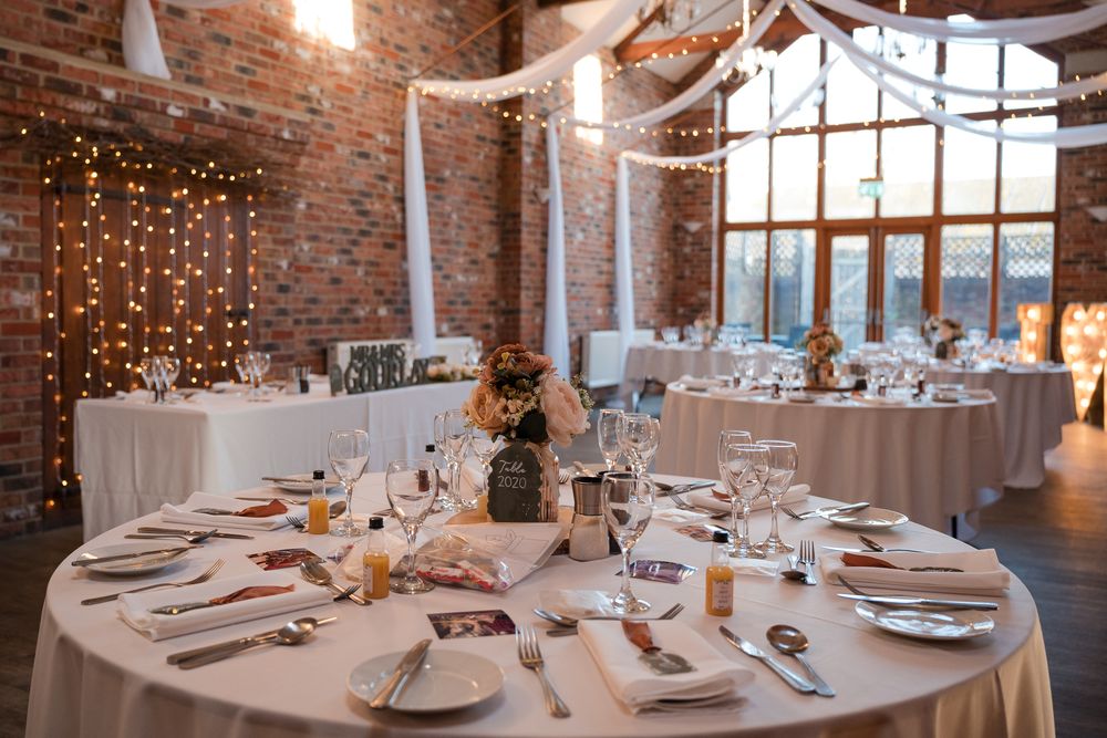 Round tables with white linens and place settings arranged in an industrial brick loft space with string lights overhead.