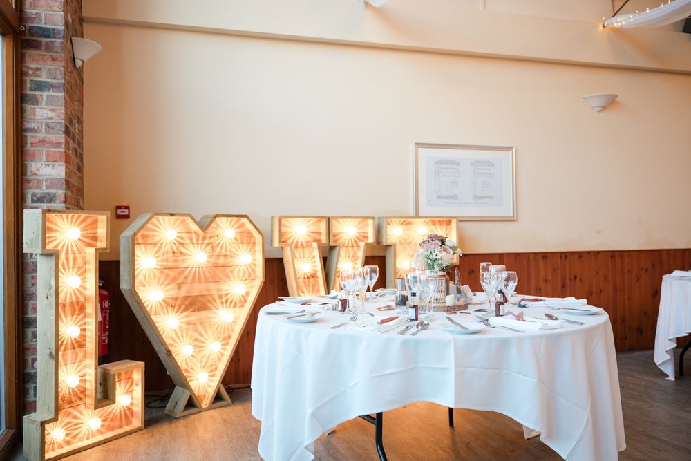Elegant wedding reception venue with illuminated LOVE marquee letters and white tablecloths in a rustic brick setting.