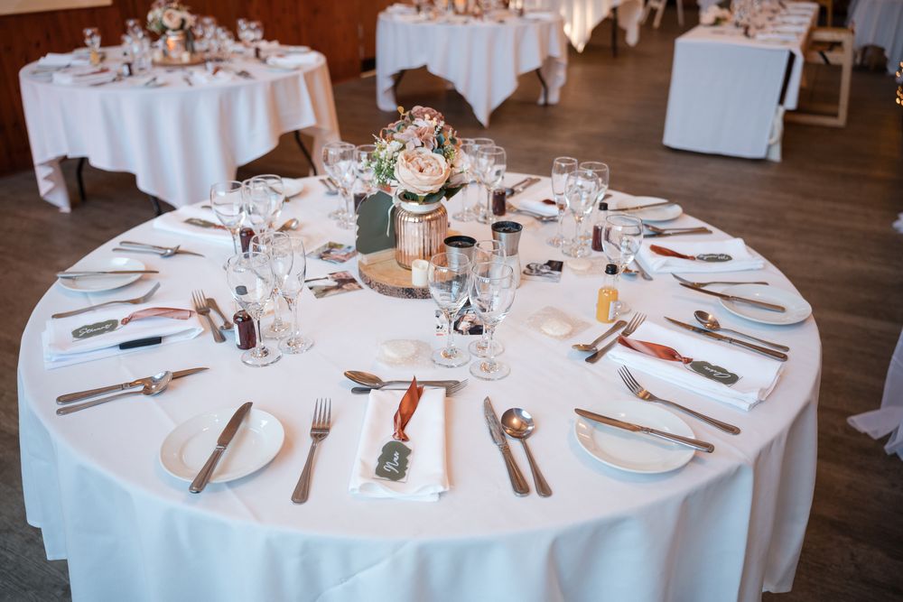 Elegant round wedding reception table with white linens, place settings, and floral centerpiece in rose gold accents.