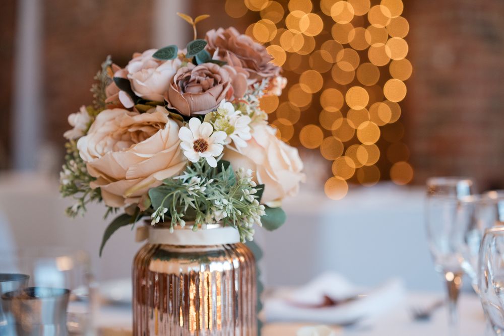 Elegant wedding reception table settings with blush flowers in mercury glass vases and warm bokeh lighting in background.