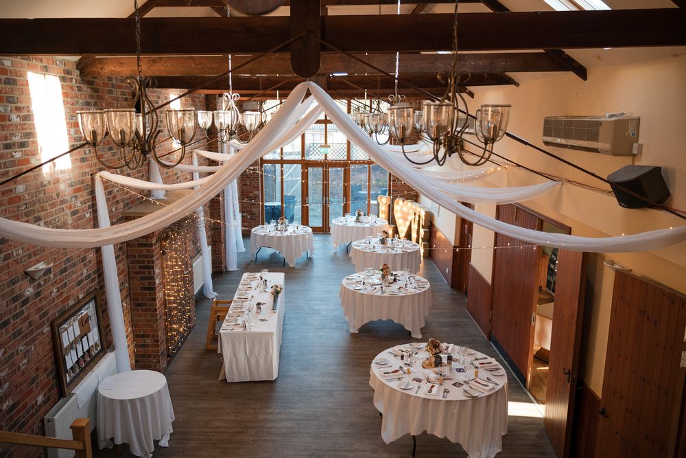Rustic wedding venue interior with exposed wooden beams, elegant chandeliers, and white-draped tables set for reception.