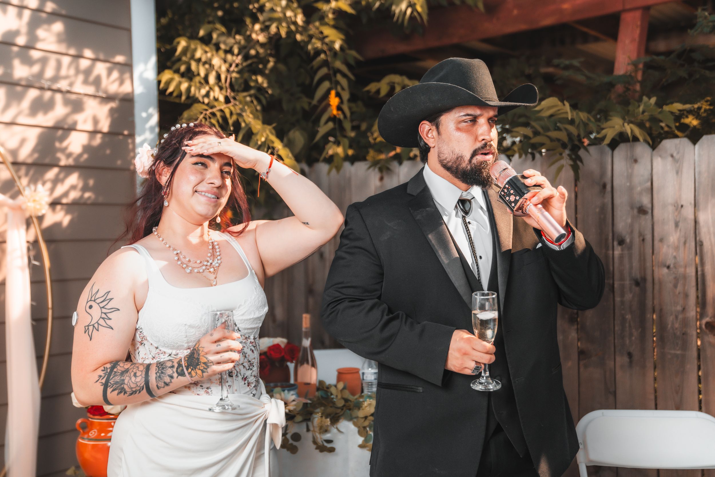 A bride and groom share a laugh while drinking from wine glasses at their outdoor wedding reception.