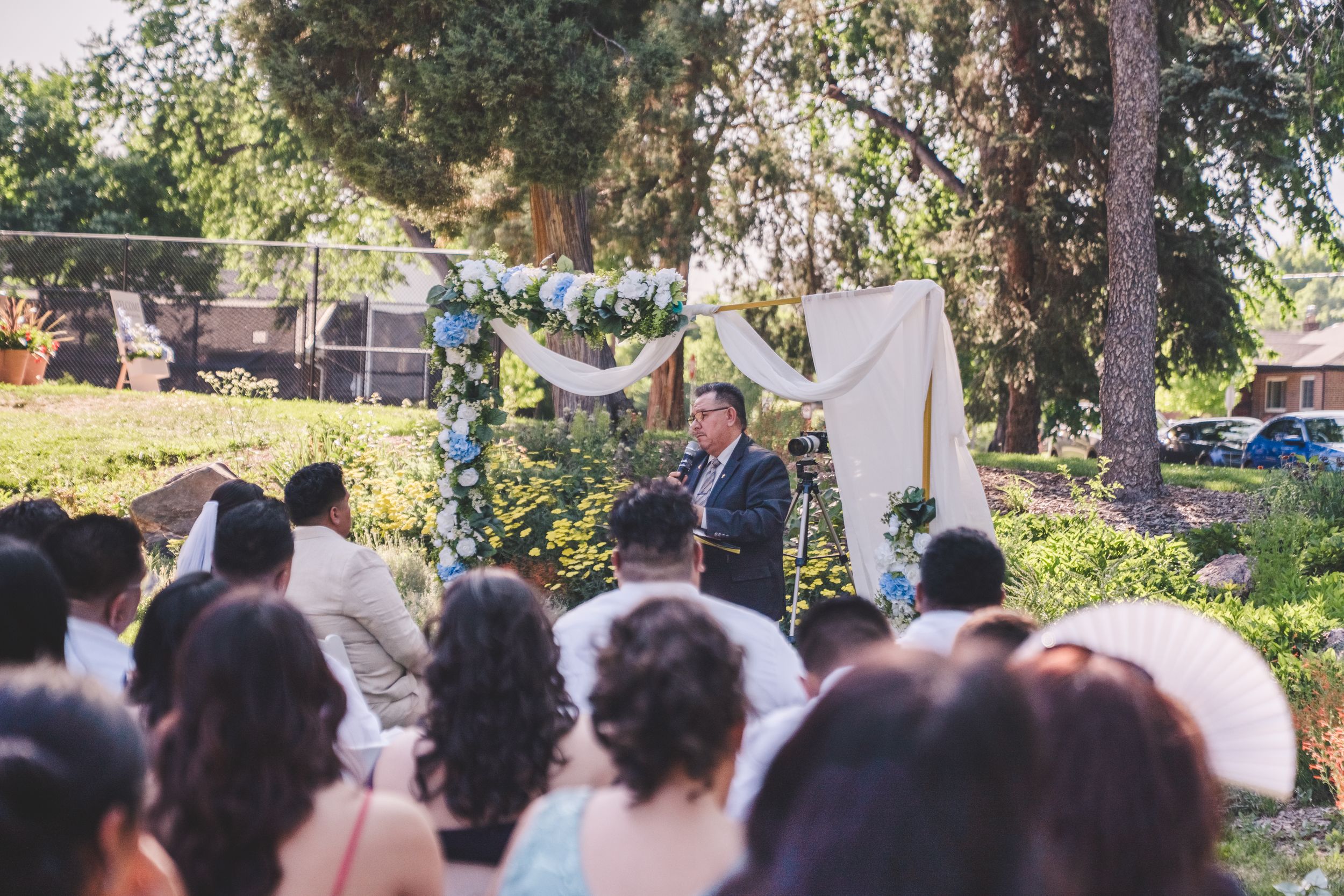 Wedding guests gather outdoors under tall trees during a sunny garden ceremony celebration.