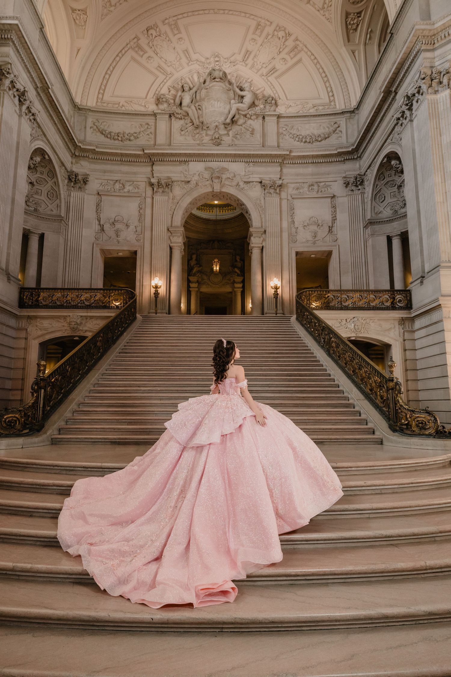 A flowing pink ballgown photographed on grand marble stairs in an ornate classical building interior.
