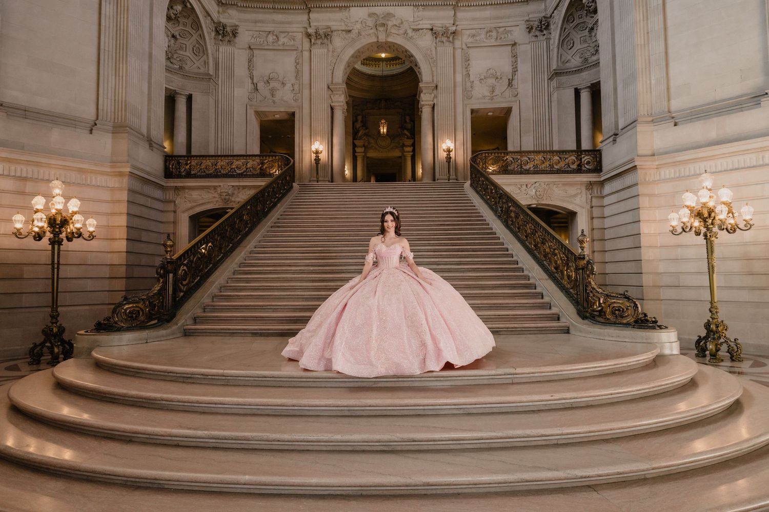 A figure in a pink ballgown poses on grand marble steps inside an ornate classical building with elegant chandeliers.
