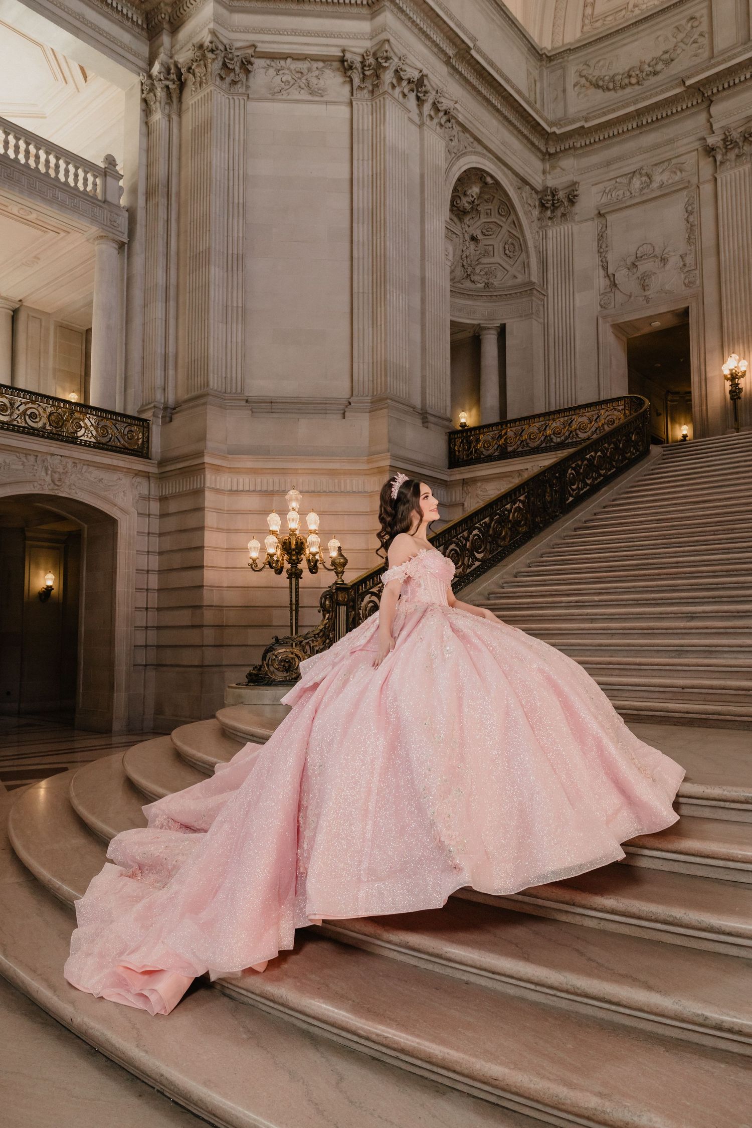 A stunning pink ballgown with a flowing train photographed on an elegant marble staircase in a grand historic building.