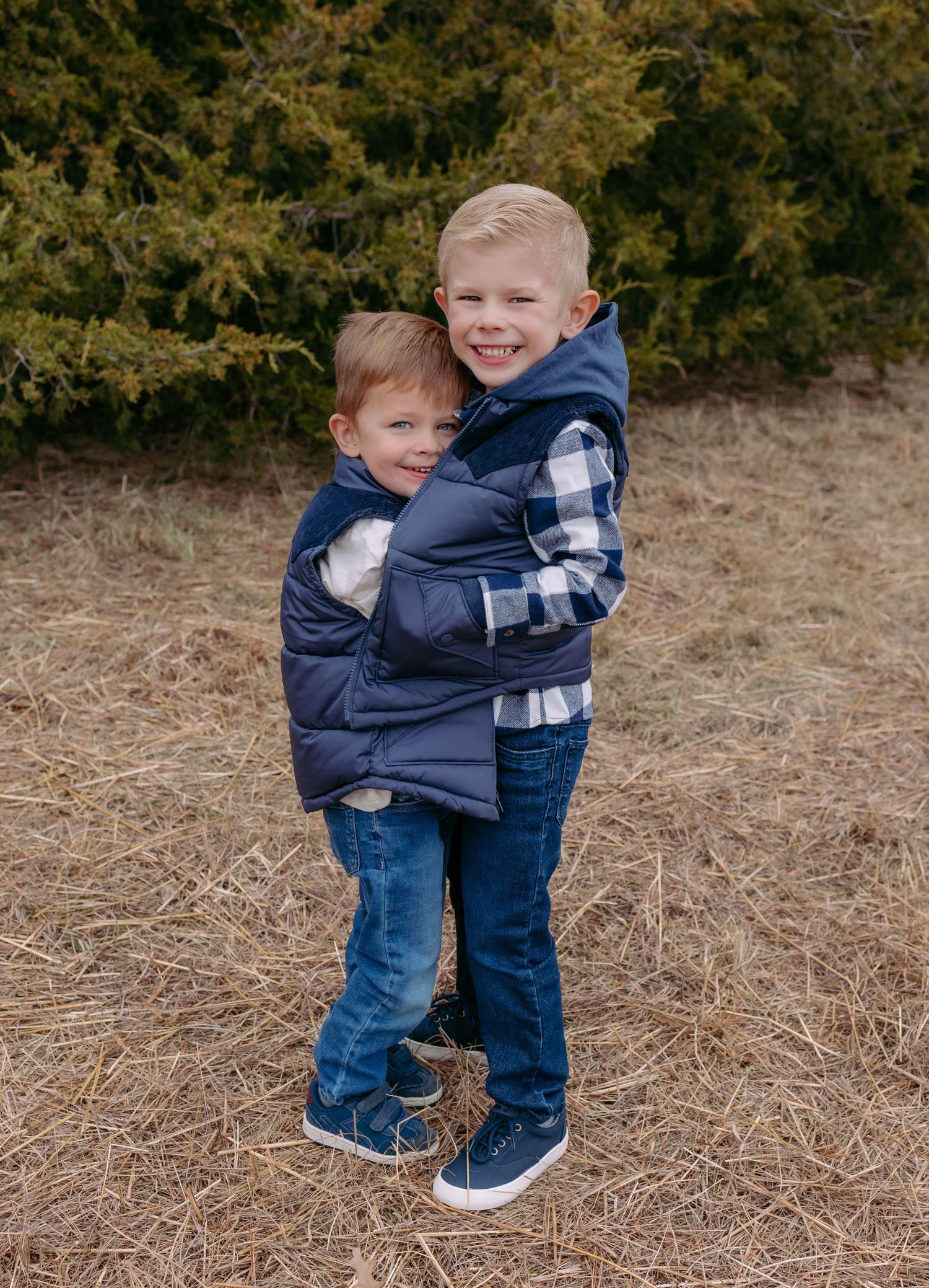 Two young siblings in blue and plaid outfits share a sweet embrace while standing in a grassy outdoor setting.