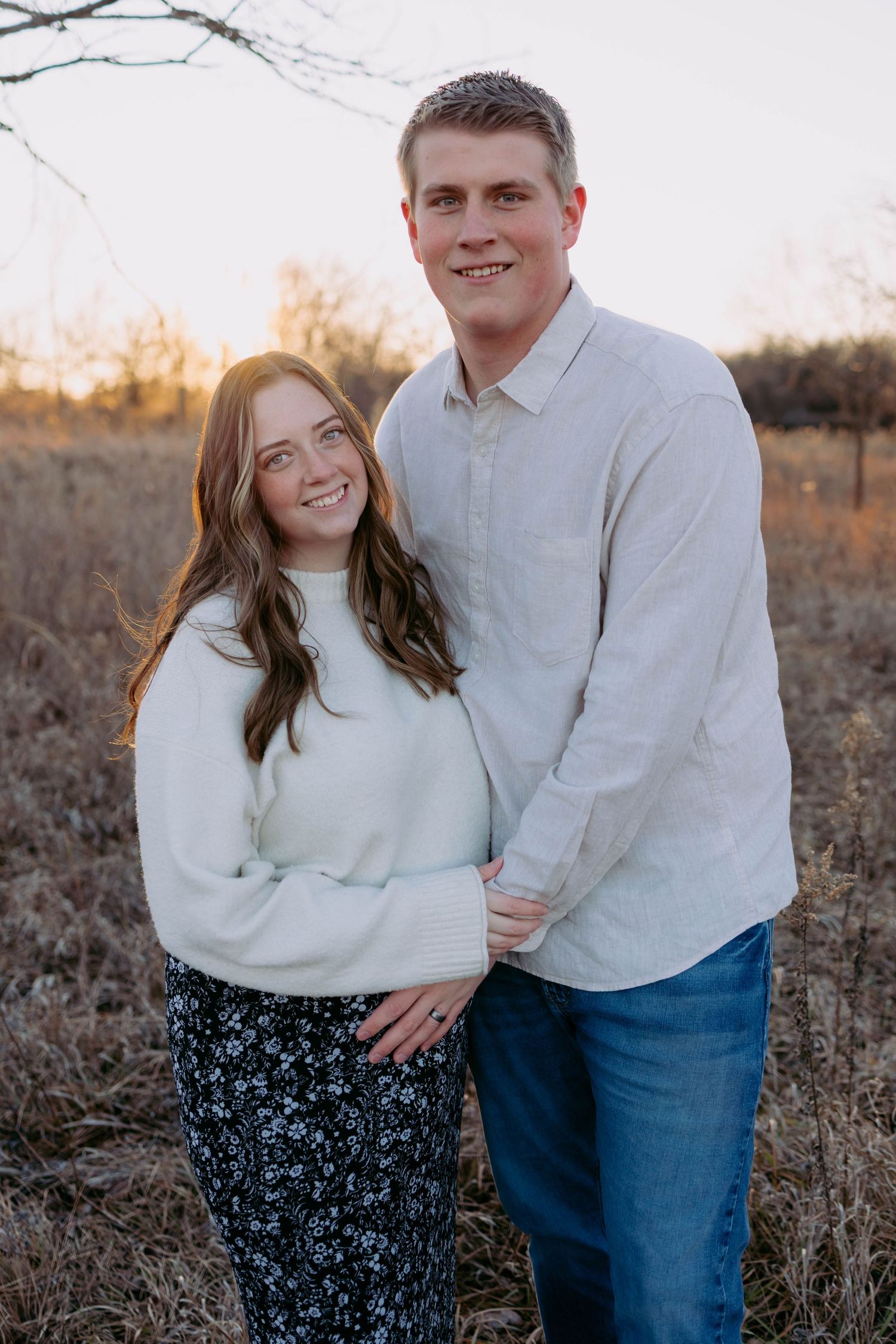 A couple in matching white sweaters share romantic moments during a sunset photoshoot in a rustic field.