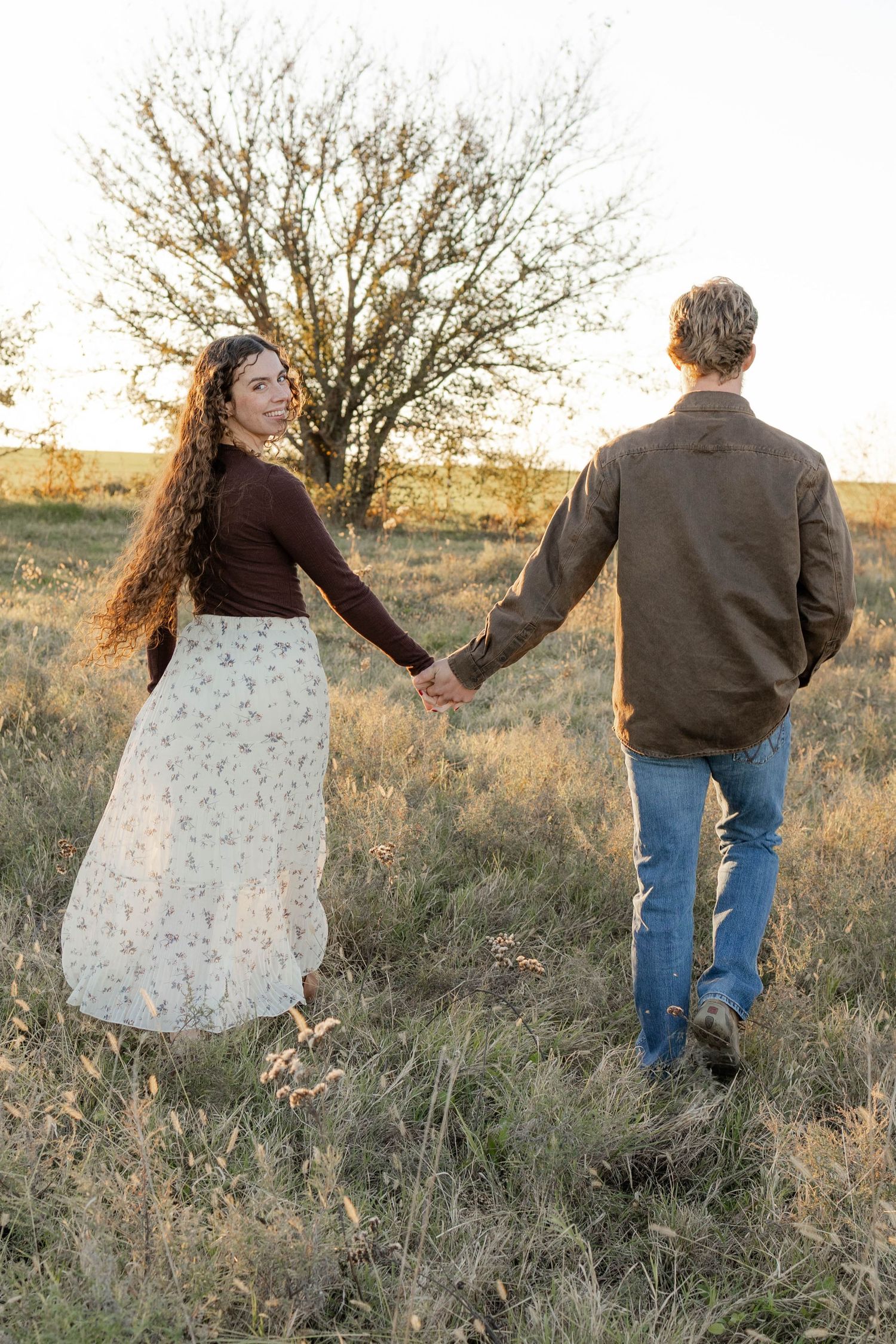 True West Photography - A couple holds hands while walking through a golden field at sunset with a bare tree in the background.