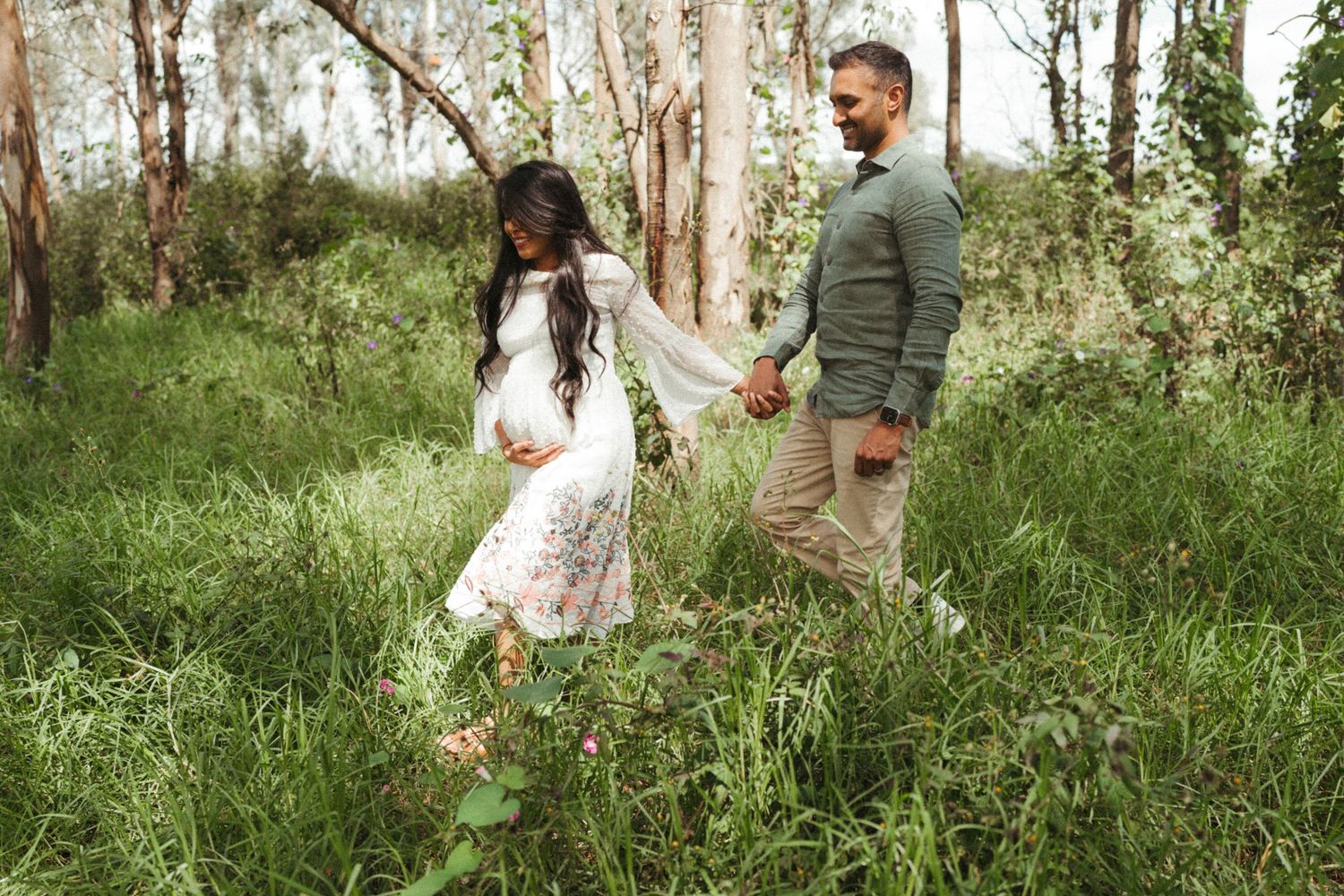 Romantic couple holding hands while walking through lush green forest setting.