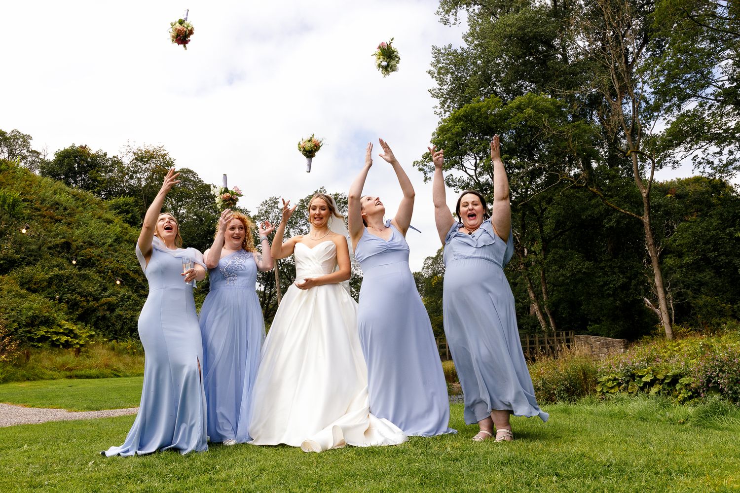 Wedding party in light blue bridesmaid dresses and white bridal gown pose joyfully on grassy hillside.