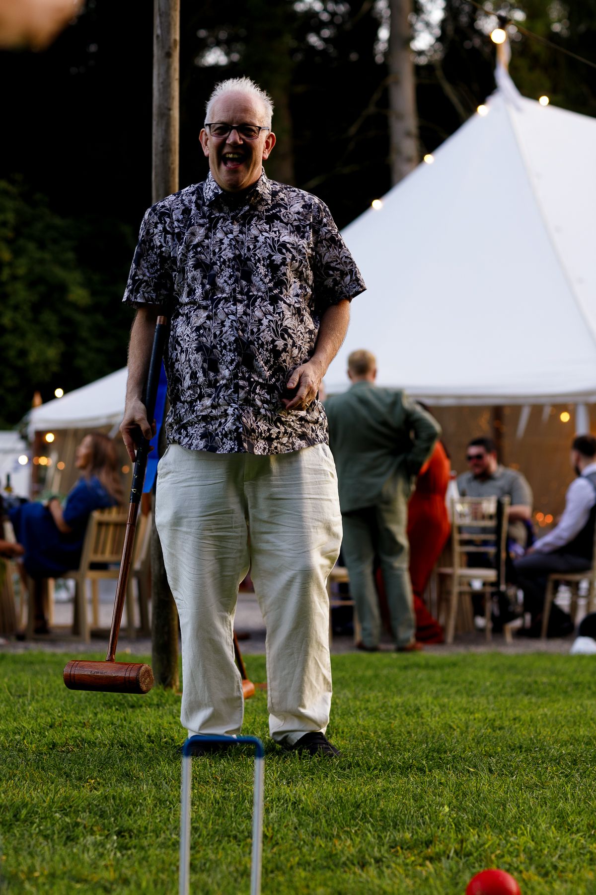 A person wearing a floral print shirt and khaki pants stands on grass near white event tents at an outdoor gathering.