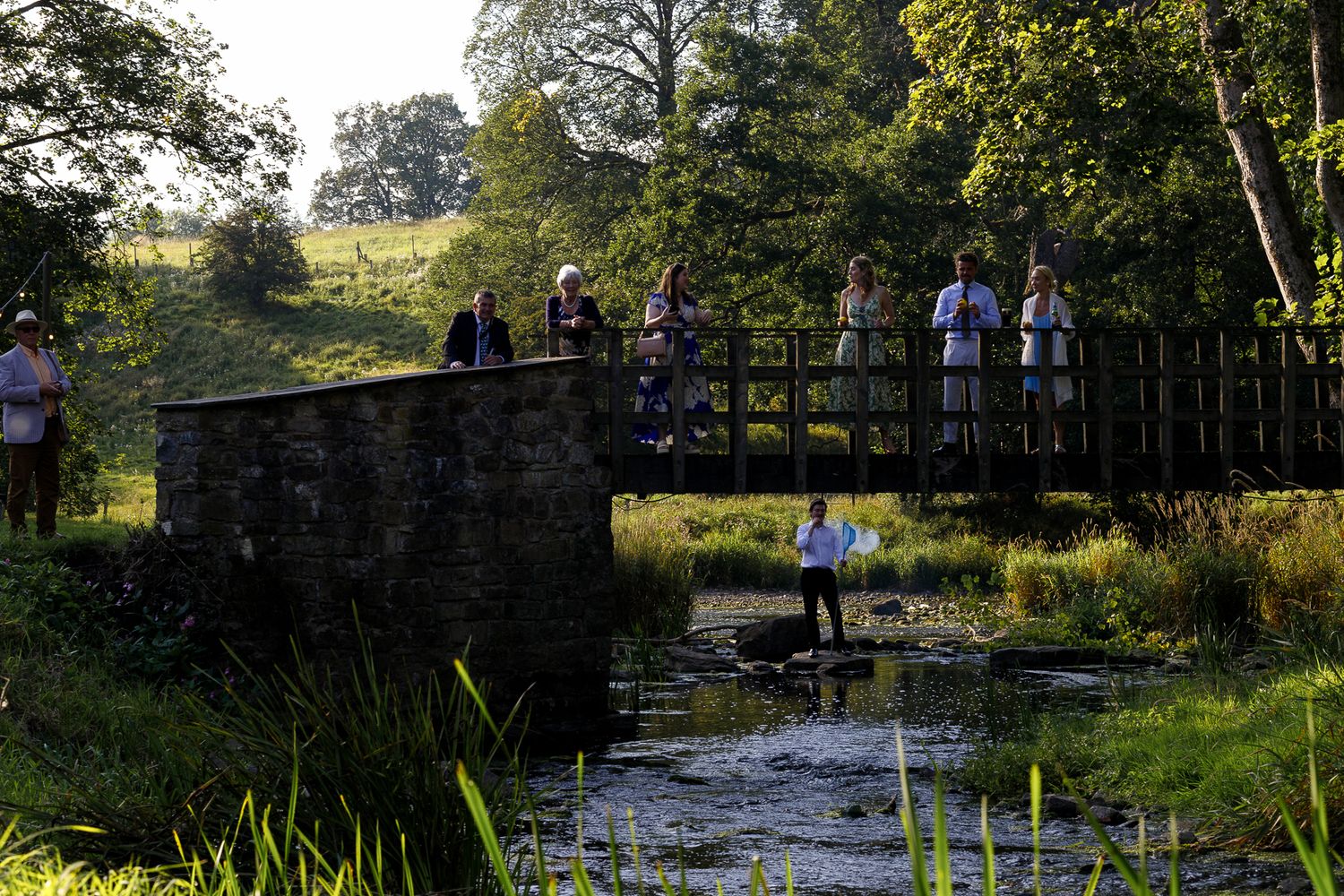 People gather on a stone bridge overlooking a tranquil stream surrounded by lush greenery at sunset.