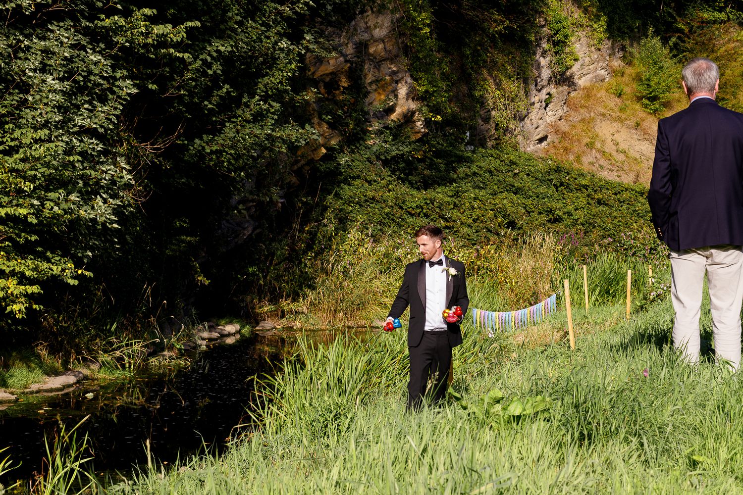 Two people stand in a grassy area near a stream with trees and rocky cliffs in the background.