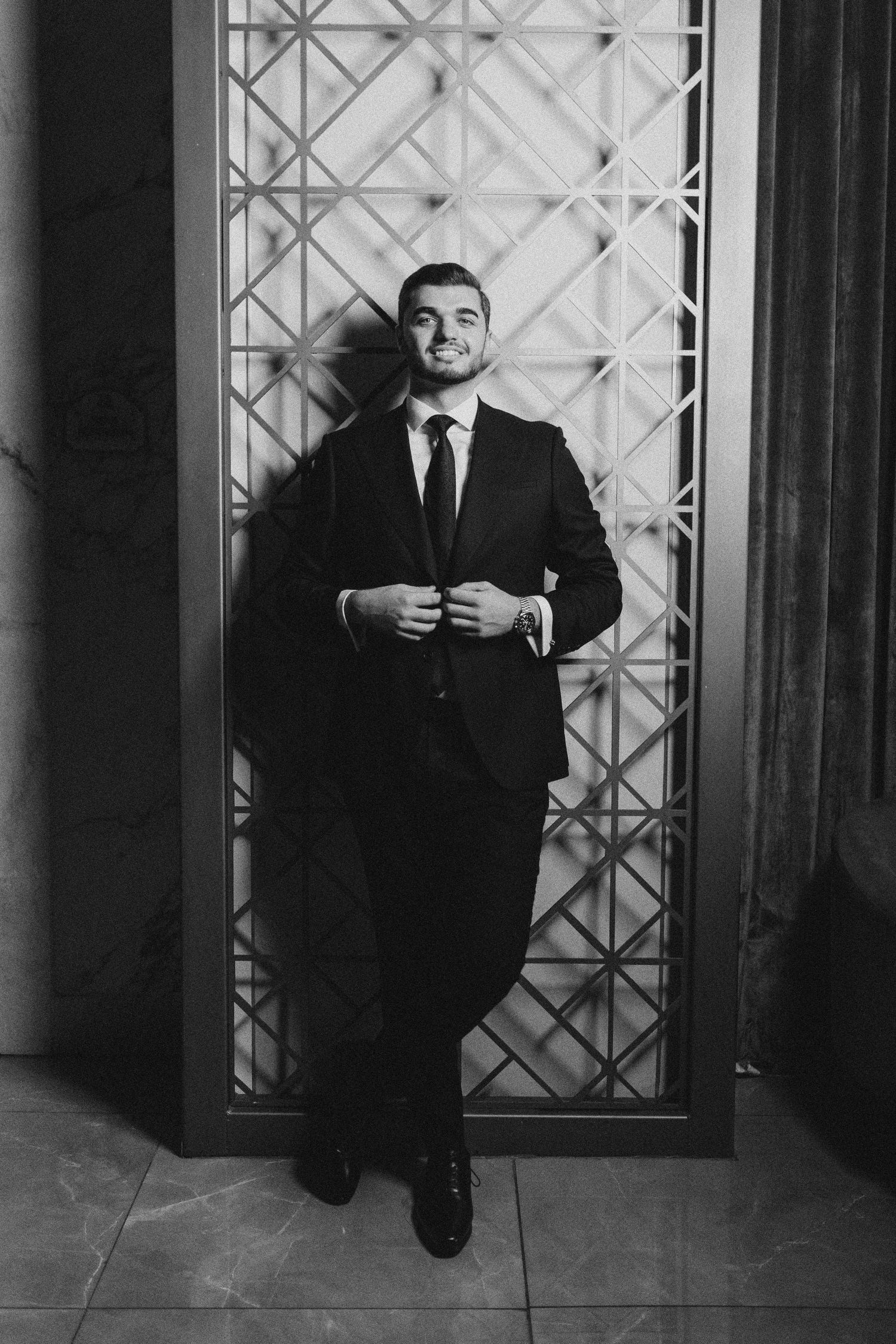 Moody black and white portraits of a gentleman in a suit against a decorative window backdrop.