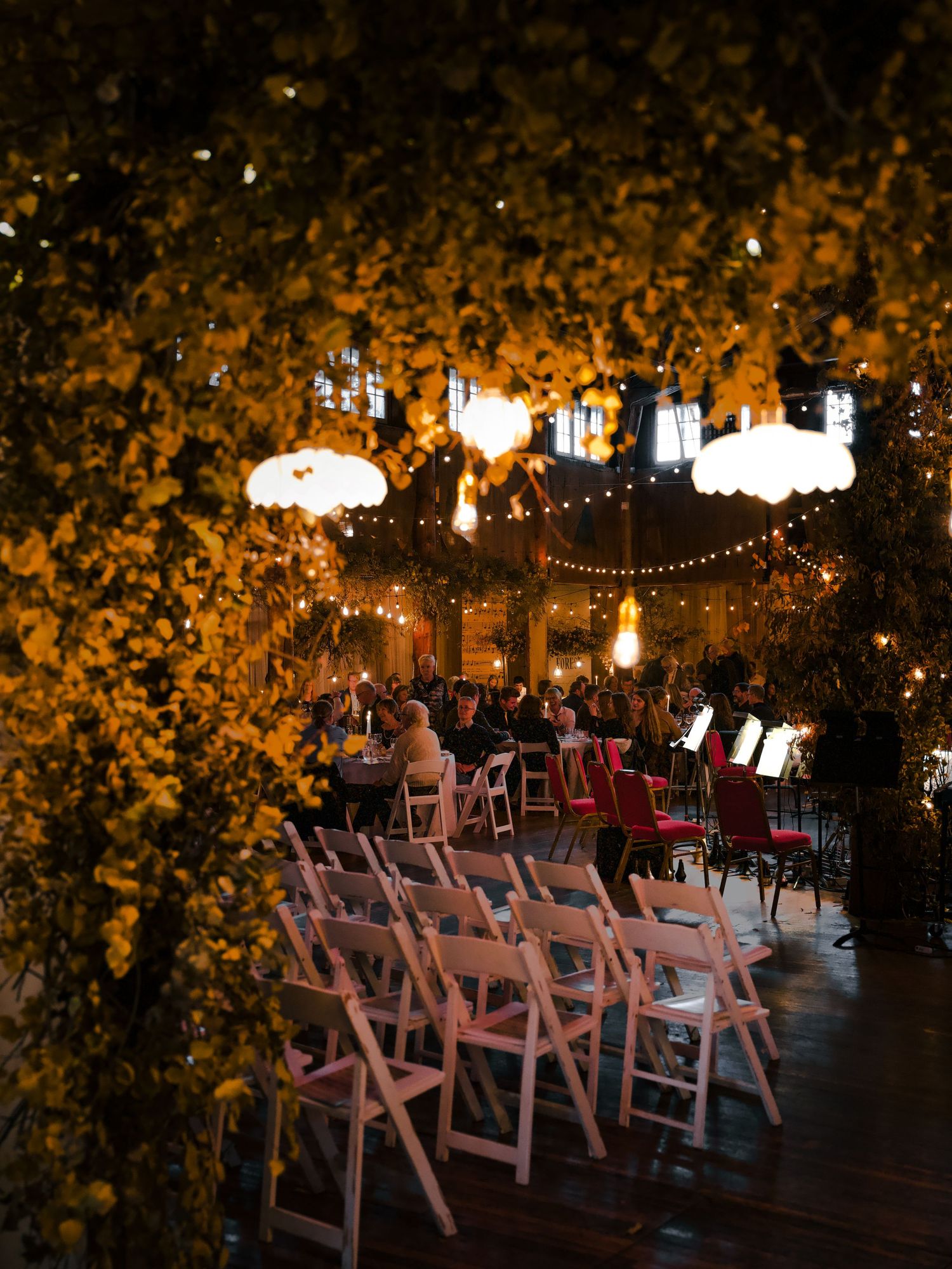 White folding chairs arranged in rows under string lights and autumn foliage.
