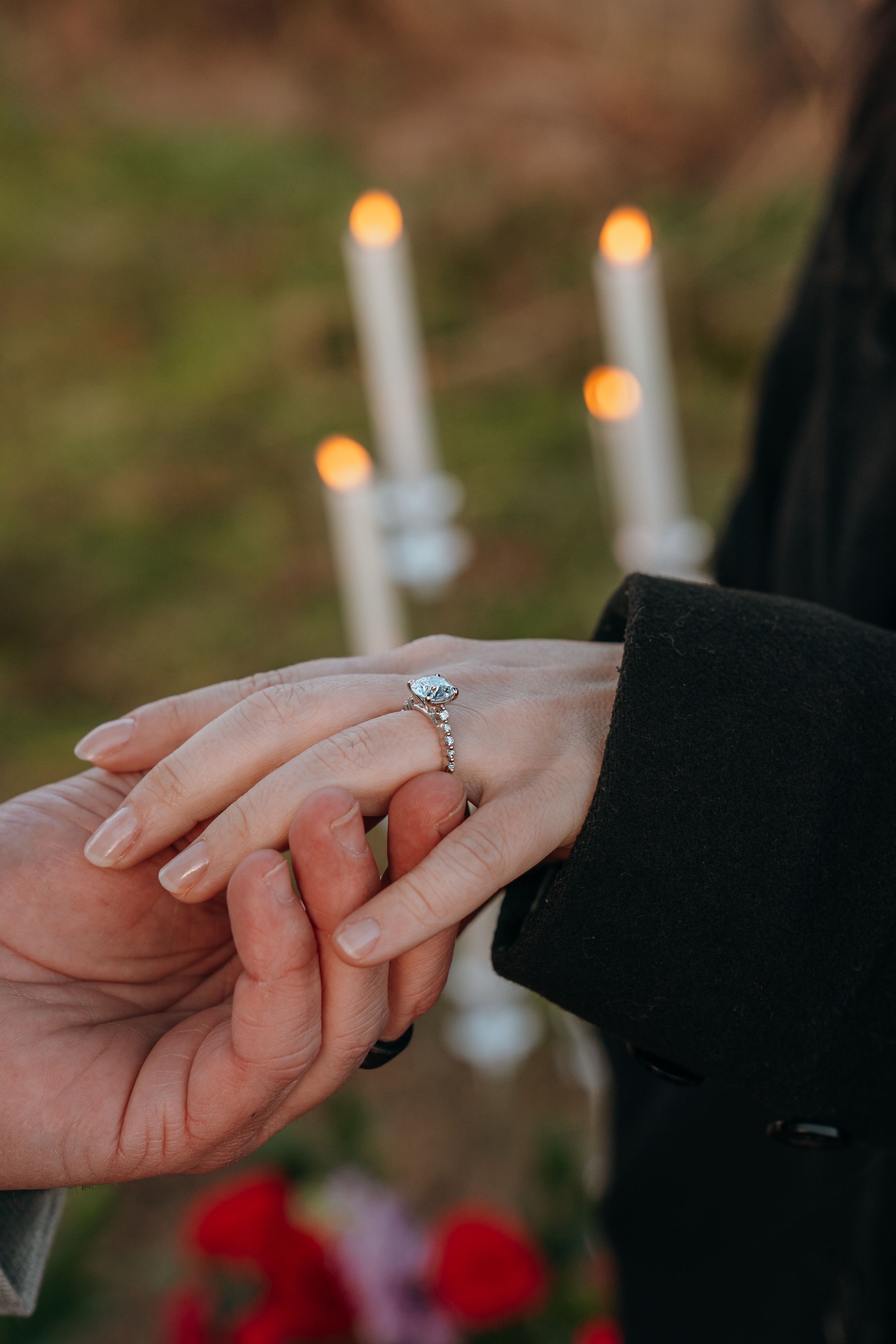 Close-up of hands showing engagement ring with lit white candles and red roses in the background.