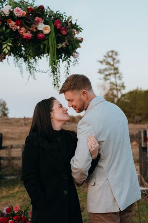A romantic moment under a floral arch with pink roses in a rustic outdoor setting at sunset.