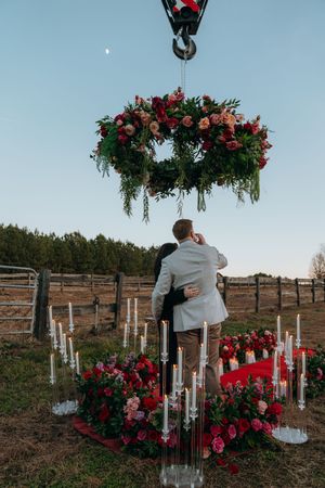 Romantic outdoor proposal setup with red roses, glass cylinders, and candles arranged on a hillside at sunset.