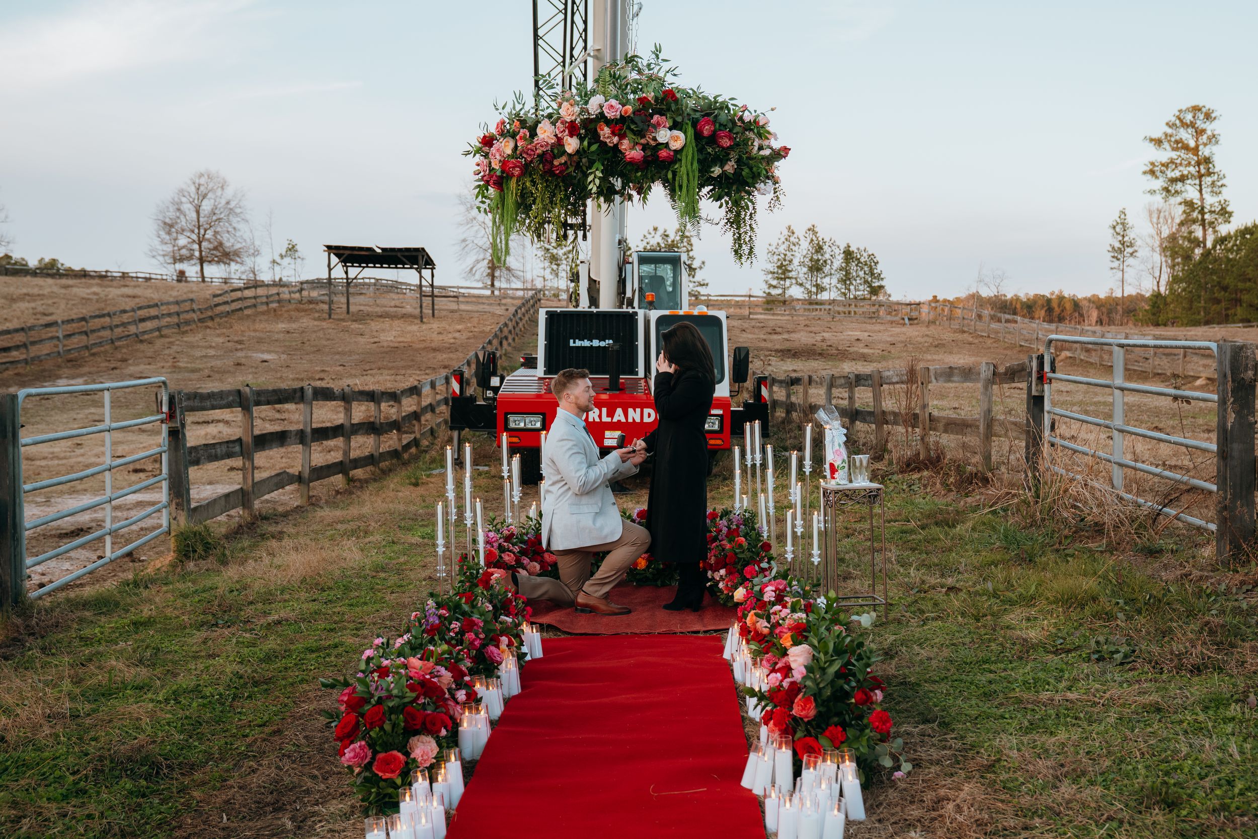 Outdoor wedding ceremony setup with red carpet aisle runner and rustic wooden arch decorated with red flowers.