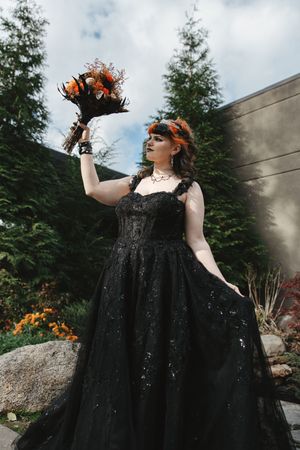 A dramatic figure in a black sparkly dress poses outdoors against evergreen trees with orange floral crown details.
