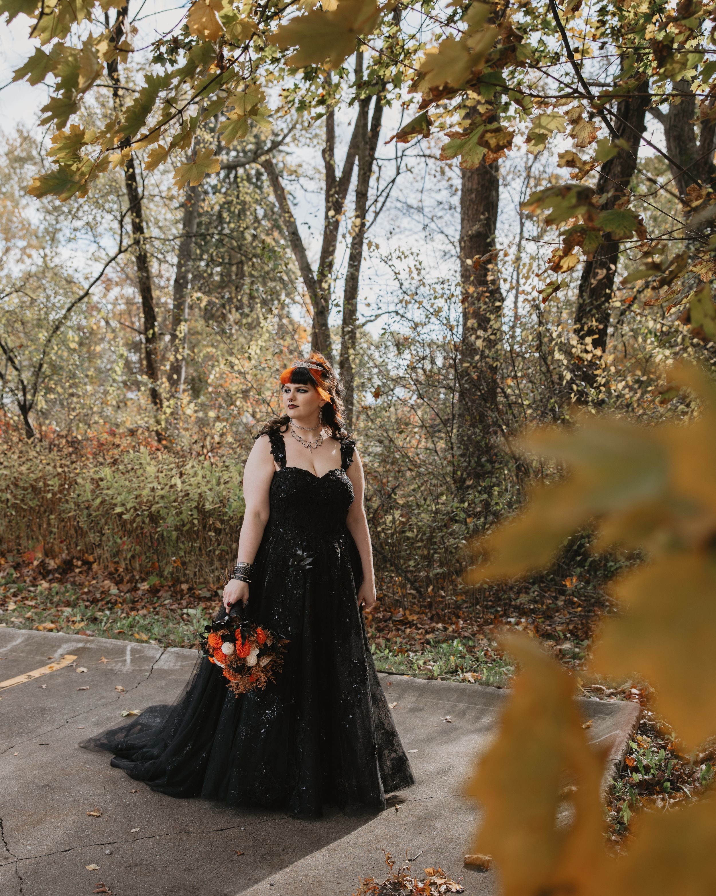 A figure in a flowing black gown poses gracefully among autumn foliage on a stone path.
