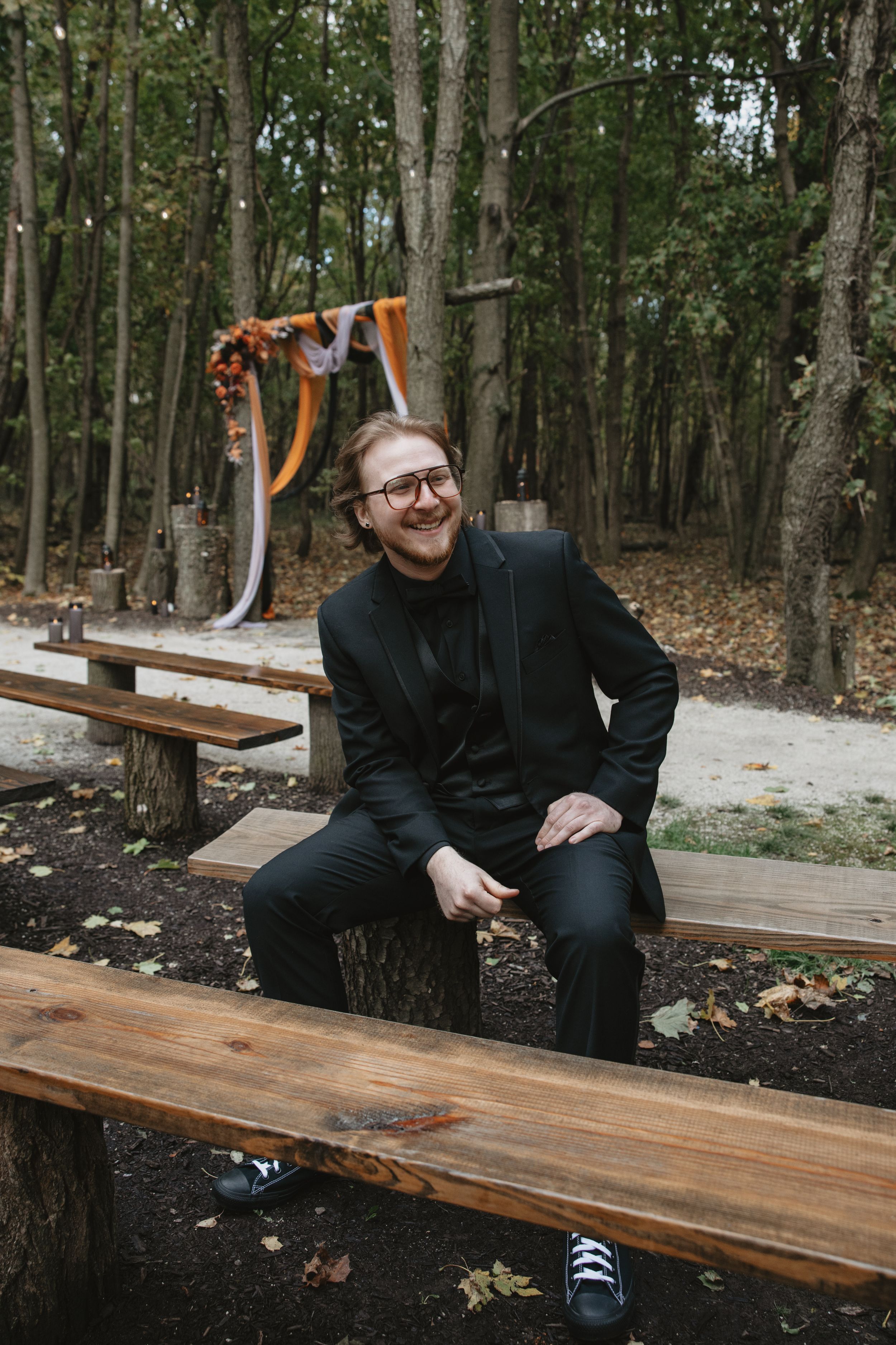 A person in dark attire sits casually on a wooden bench in a wooded outdoor setting.