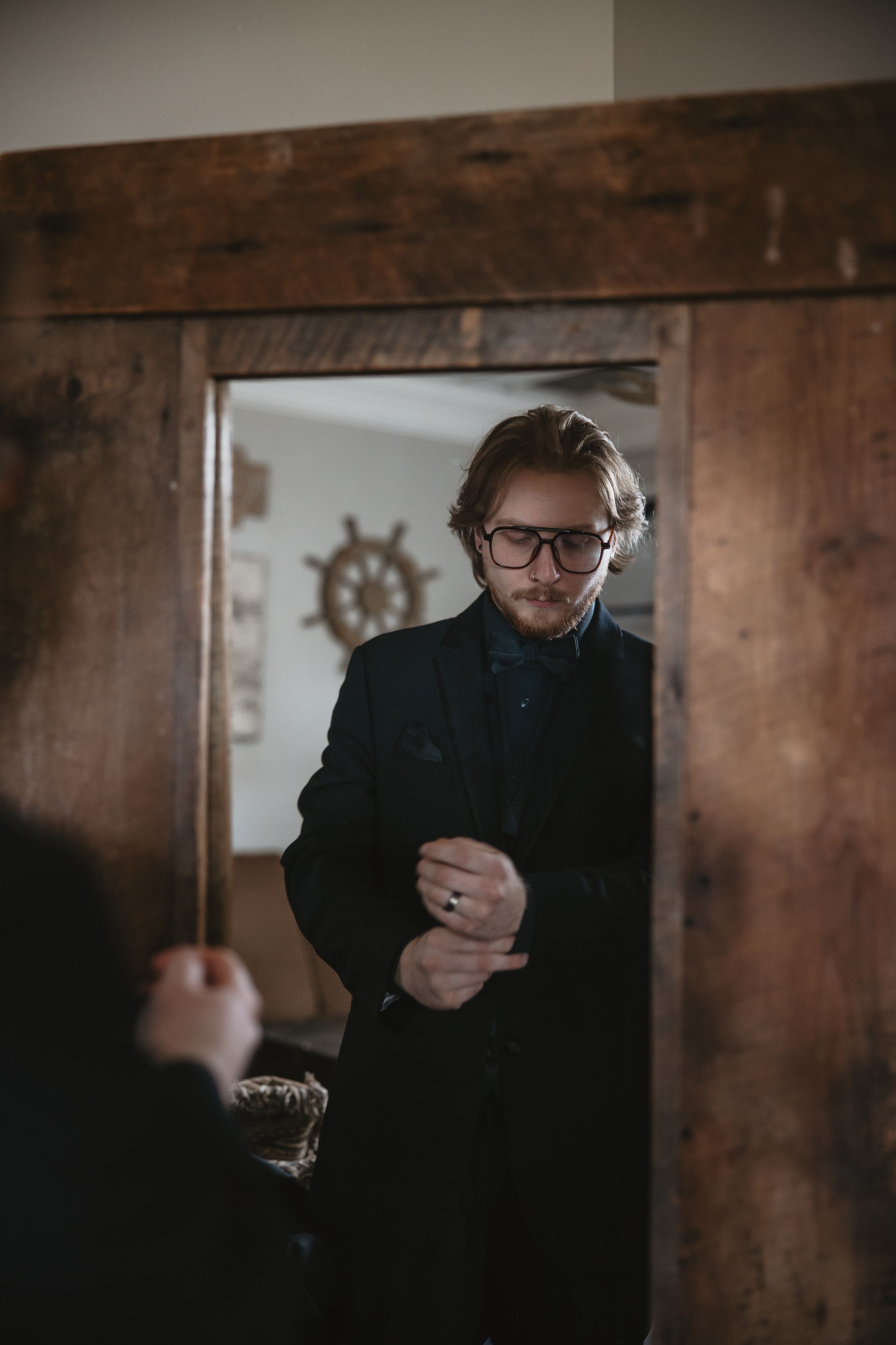 A moody portrait in dark tones showing a person in black attire and glasses adjusting their sleeve by a wooden doorway.